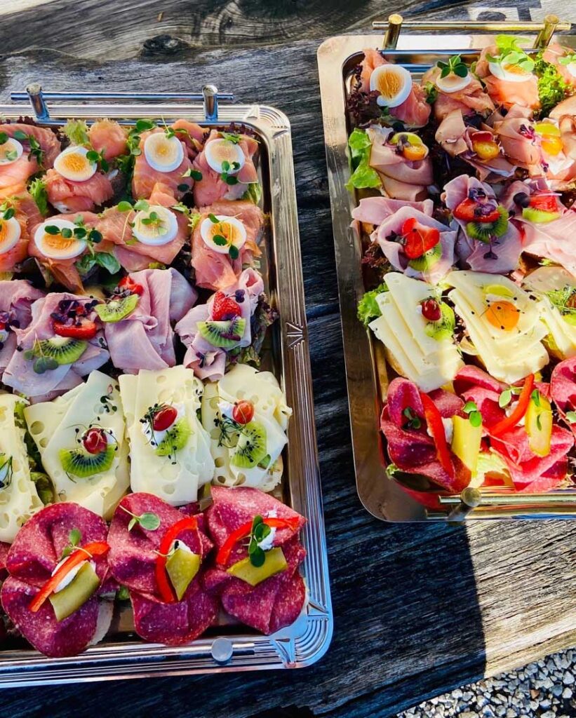 Zwei Metalltabletts mit verschiedenen belegten Brötchen - belegt mit Fleisch, Käse, Eiern, Kiwi, Essiggurken, Tomaten und Kräutern - werden auf einem rustikalen Holztisch in hellem Sonnenlicht wunderschön präsentiert, perfekt für jedes Catering oder jeden Partyservice.