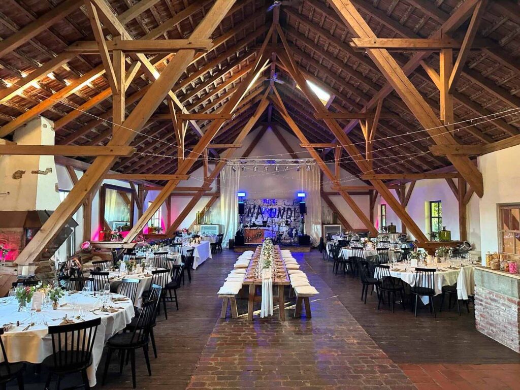 Eine rustikale, für eine Hochzeit dekorierte Scheune mit Holzbalken, Lichterketten, langen Banketttischen mit weißen Tüchern und Blumenschmuck sowie einem Firmencatering, das leckere Köstlichkeiten unter einem beleuchteten "LOVE"-Schild an der Rückwand serviert.
