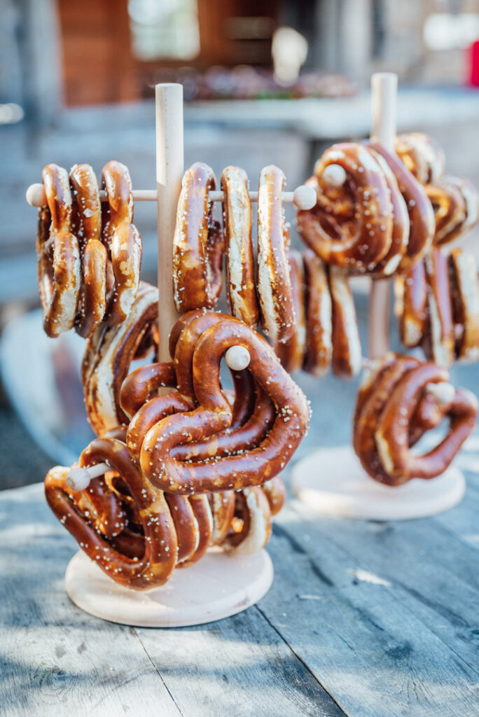 Holzständer halten große, gesalzene Brezeln, einige in Herzform, die im Freien auf einem rustikalen Holztisch präsentiert werden - perfekt für ein einzigartiges Hochzeitscatering oder ein stilvolles Firmencatering in hellem Sonnenlicht.