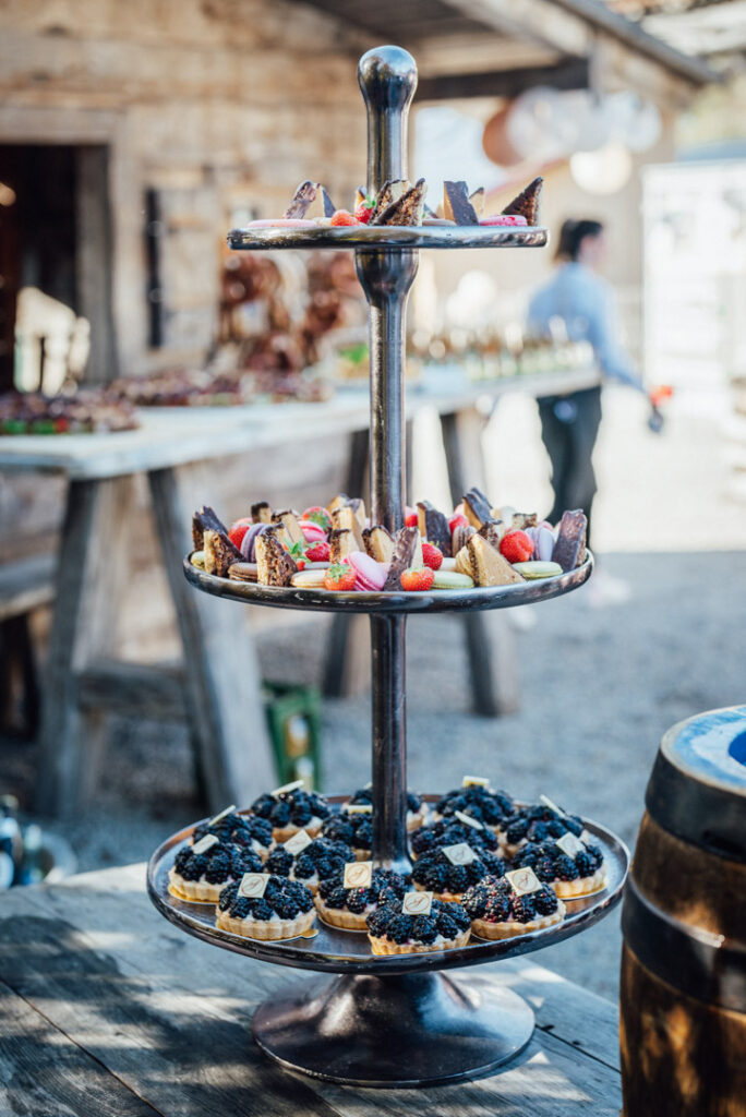 Ein dreistöckiger Dessertstand mit verschiedenen Kuchen und Torten, belegt mit Beeren und Schokolade, steht im Freien auf einem rustikalen Holztisch - perfekt für Catering-Veranstaltungen oder Firmencatering in Passau.