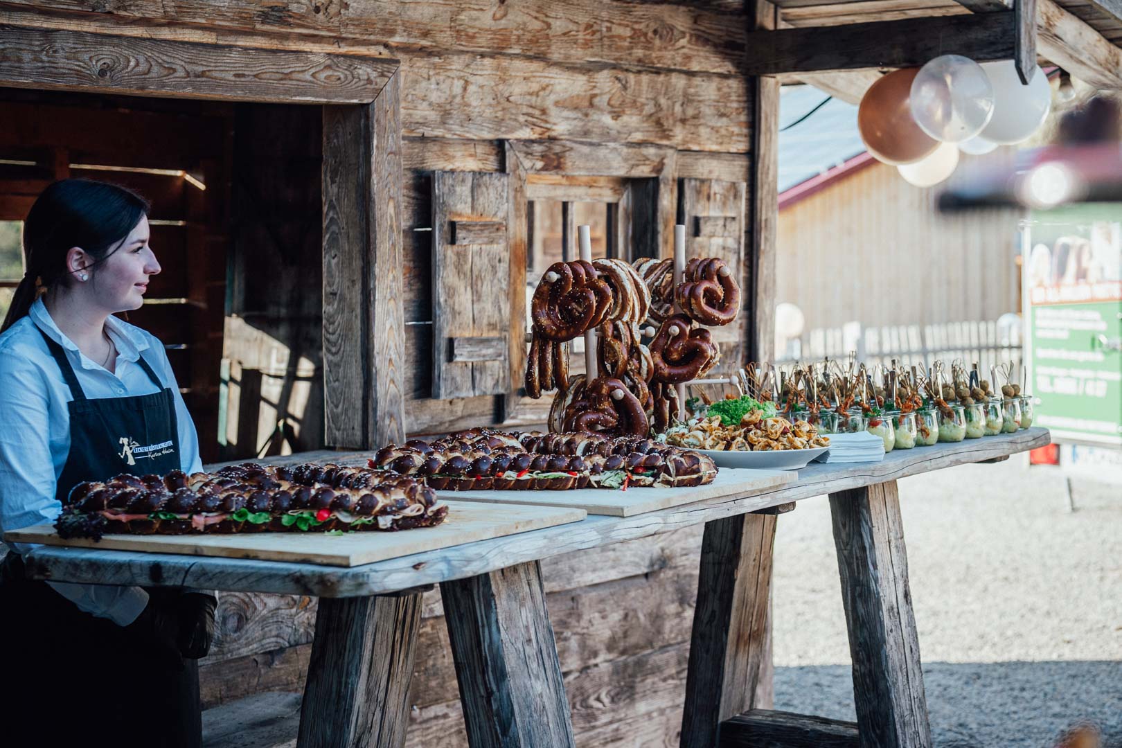 Eine Frau in schwarzer Schürze steht neben rustikalen Holztischen mit verschiedenen Brezeln, Gebäck, Snacks und Häppchen - perfekt für das Hochzeitscatering oder den Partyservice in Passau - vor einem Holzgebäude mit Luftballons auf dem Dach.