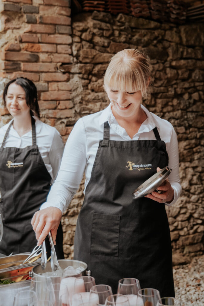 Zwei Frauen von Karina Haim, Leckere Köstlichkeiten Catering Passau, gekleidet in schwarze Schürzen und weiße Hemden, stehen neben einem Tisch mit Gläsern und Speisen. Eine hält lächelnd eine Zange und ein Klemmbrett in der Hand, die andere steht vor einer rustikalen Steinmauer.