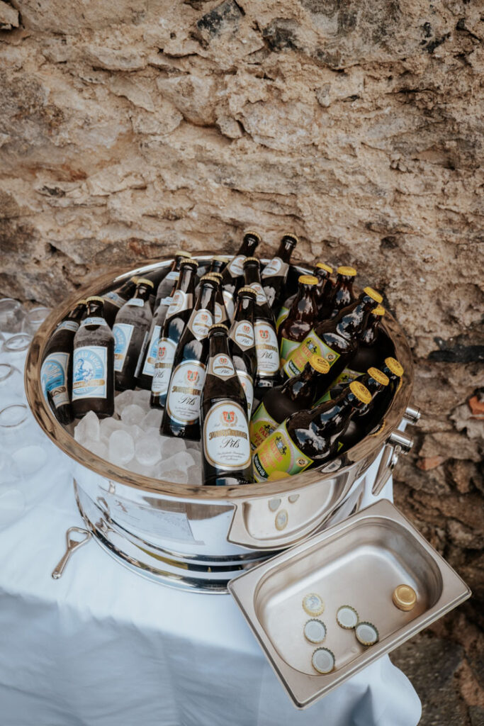 Eine große Metallschüssel, gefüllt mit Eis und verschiedenen Bierflaschen, steht auf einer weißen Tischdecke neben einer Steinwand - perfekt für Partyservice oder Hochzeitscatering. Mehrere Bierflaschenverschlüsse liegen in einem kleinen Metalltablett daneben.