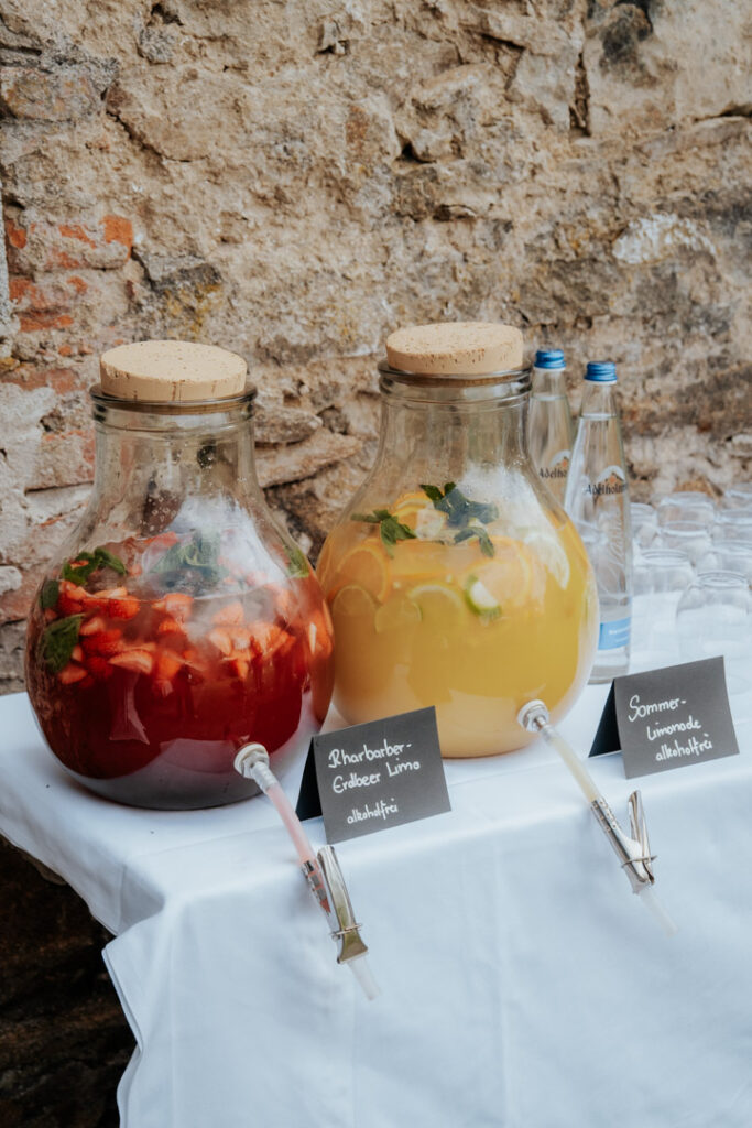 Perfekt für das Hochzeitscatering Passau oder das Partyservice: Auf einem weißen Tisch stehen zwei große gläserne Getränkespender mit Korkdeckeln; einer enthält rote Fruchtbowle mit Erdbeeren und Kräutern, der andere gelbe Bowle mit Zitrone und Minze. Kreideschilder beschriften jedes Getränk.