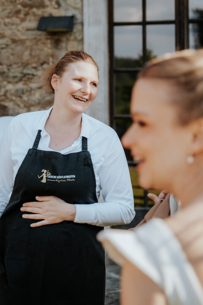 Karina Haim, von Leckere Köstlichkeiten Catering Passau steht draußen in der Nähe eines Steingebäudes, während eine andere Frau in einem weißen Kleid im Vordergrund unscharf zu sehen ist.