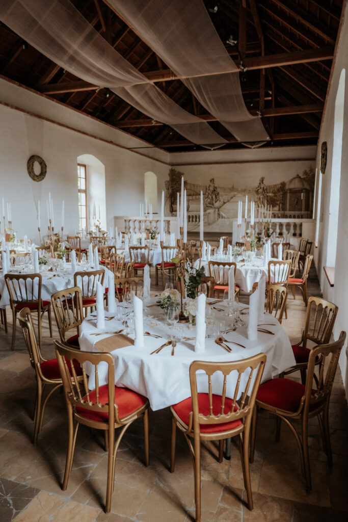 Ein dekorierter Festsaal in Passau bietet runde Tische mit weißen Tüchern, eleganten Tellern, Gläsern, hohen weißen Kerzen und schlichten Blumensträußen - ideal für Veranstaltungen mit professionellem Catering Passau. Holzstühle mit roten Sitzen sorgen für einen klassischen Touch.
