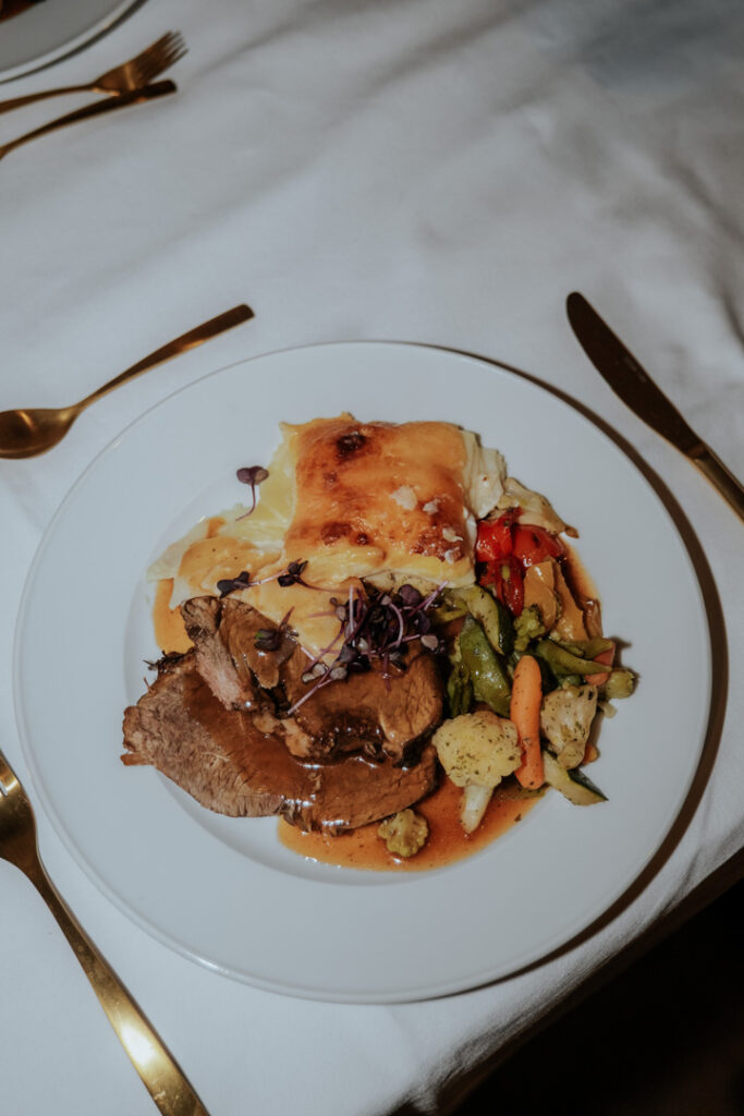 Ein weißer Teller mit Roastbeef in brauner Bratensoße, überbackenen Käsekartoffeln und einer Mischung aus gebratenem Gemüse auf einer weißen Tischdecke mit goldenem Besteck - perfekt für Catering Passau oder Ihre Hochzeitsfeier.