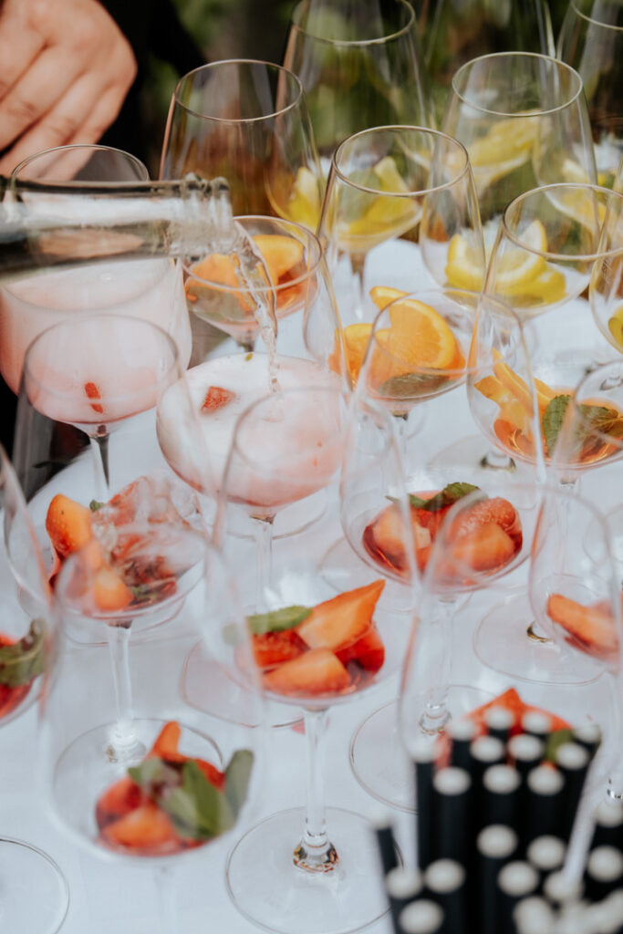 Eine Person gießt Sekt in Gläser, die mit frischen Erdbeeren und Minze gefüllt sind, perfekt für eine festliche Veranstaltung des Catering Passau. In anderen Gläsern auf dem Tisch liegen Orangen- und Limettenscheiben, im Vordergrund sind schwarze Strohhalme zu sehen.