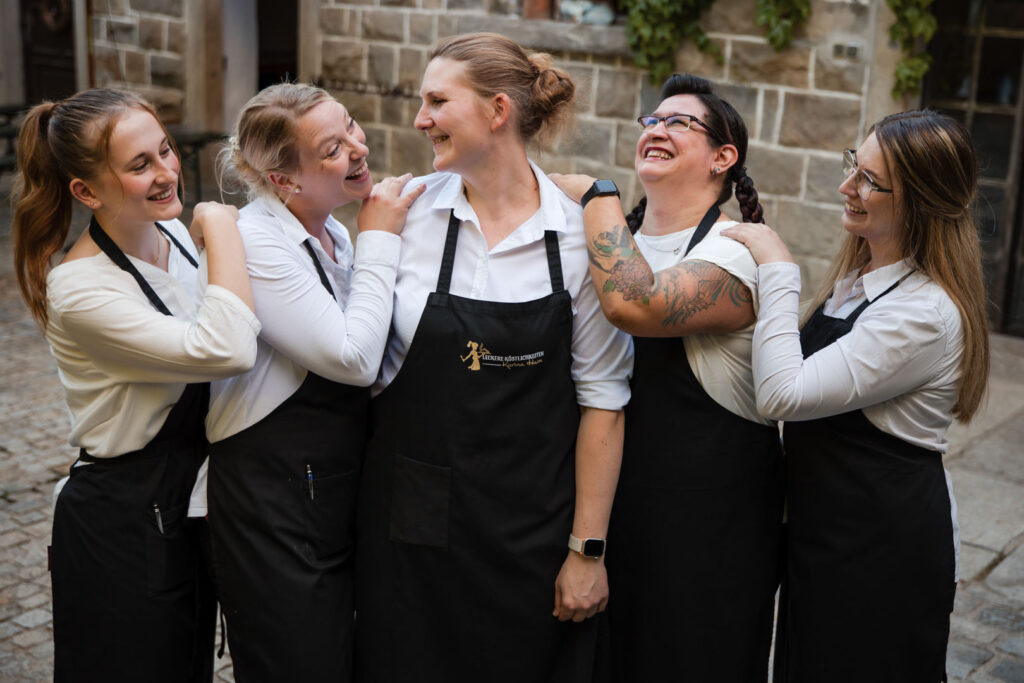 Fünf Frauen in weißen Hemden und schwarzen Schürzen stehen dicht beieinander und posieren lächelnd und lachend mit umschlungenen Armen vor einem steinernen Gebäude - Teamgeist bei Catering Passau, Firma Leckerer Köstlichkeiten.