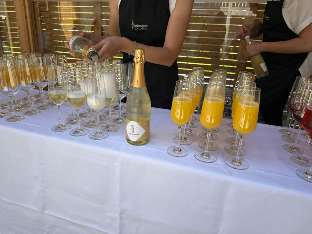 Eine Person gießt Sekt in Gläser auf einem mit weißem Tischtuch bedeckten Tisch ein, auf dem Reihen von gefüllten und leeren Gläsern stehen, darunter auch einige mit Orangensaft. Eine andere Person steht in der Nähe und hält eine Flasche.