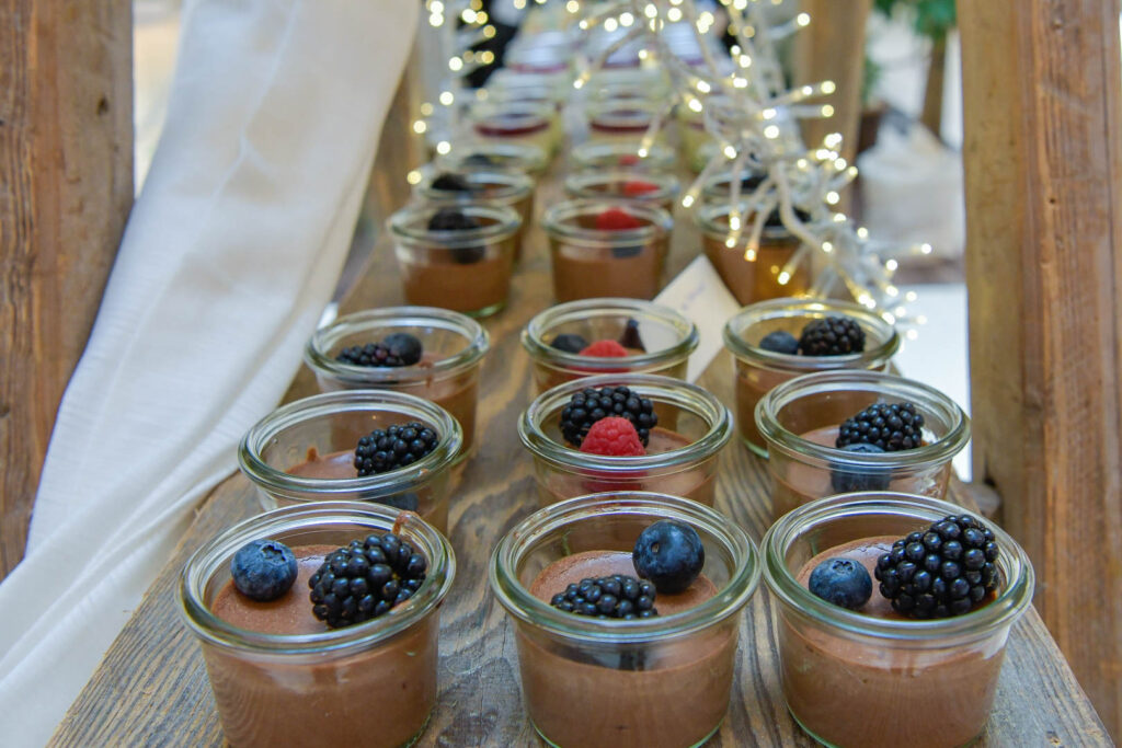 Kleine Gläser mit Mousse au Chocolat, gefüllt mit frischen Brombeeren, Heidelbeeren und Himbeeren, stehen auf einem rustikalen Holztisch - wie geschaffen für das Firmencatering in Passau, akzentuiert durch dekorative Lichterketten und ein weißes Tuch.