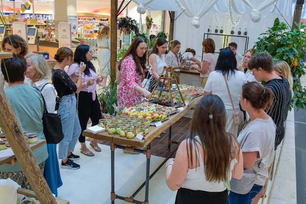 Eine Gruppe von Menschen versammelt sich bei einer Firmencatering-Veranstaltung in Passau um einen Tisch mit verschiedenen Vorspeisen und Snacks. Einige wählen das Essen aus, während andere sich unterhalten und beobachten, was eine lebhafte und gesellige Atmosphäre schafft.
