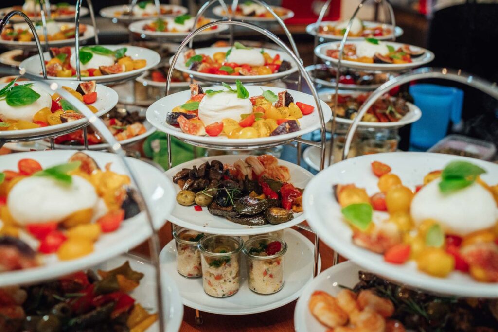 Mehrere dreistöckige Serviertabletts, gefüllt mit verschiedenen Vorspeisen - Grurrata, Tomaten, Oliven, gebratenem Gemüse und Schichtsalaten - sind für ein Catering Passau oder ein Partyservice-Buffet schön angerichtet.