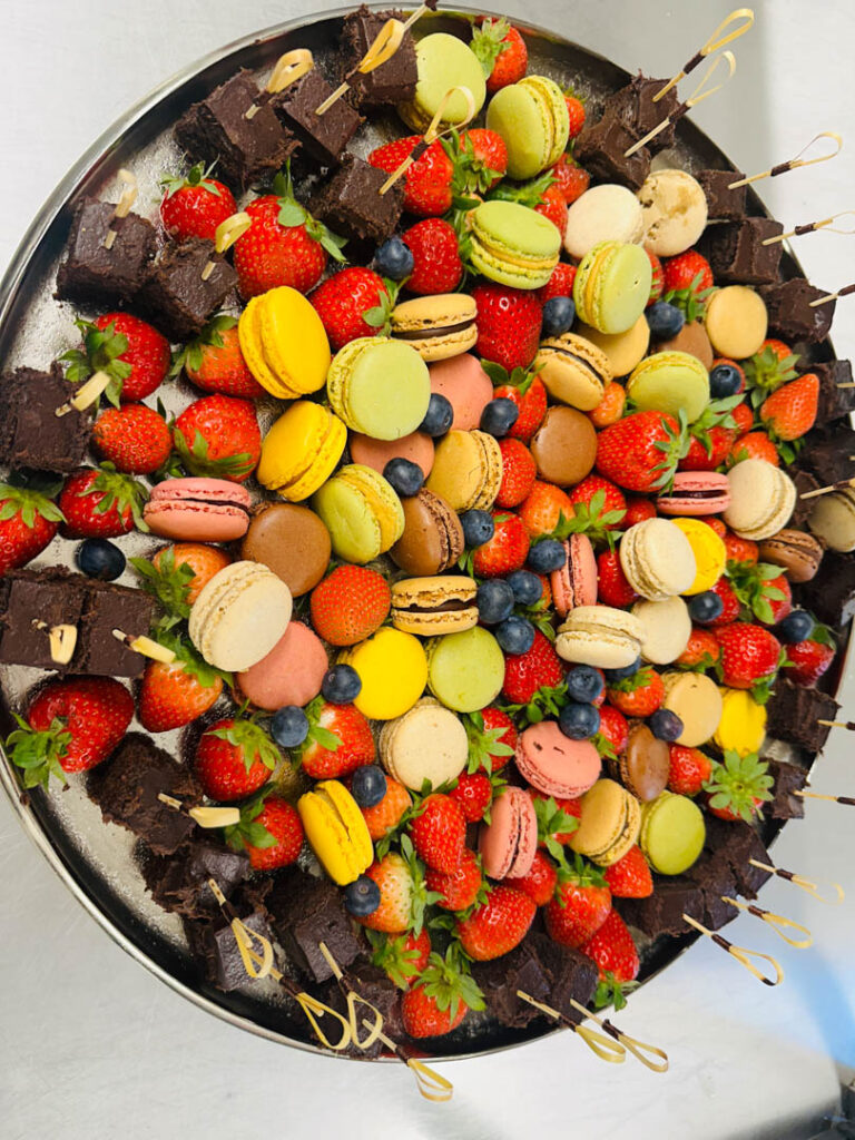 Eine große Platte gefüllt mit bunten Macarons, ganzen Erdbeeren, Blaubeeren und Schokoladen-Brownie-Häppchen, arrangiert in einem optisch ansprechenden, festlichen Display - perfekt für Catering Passau oder jede Partyservice-Veranstaltung.