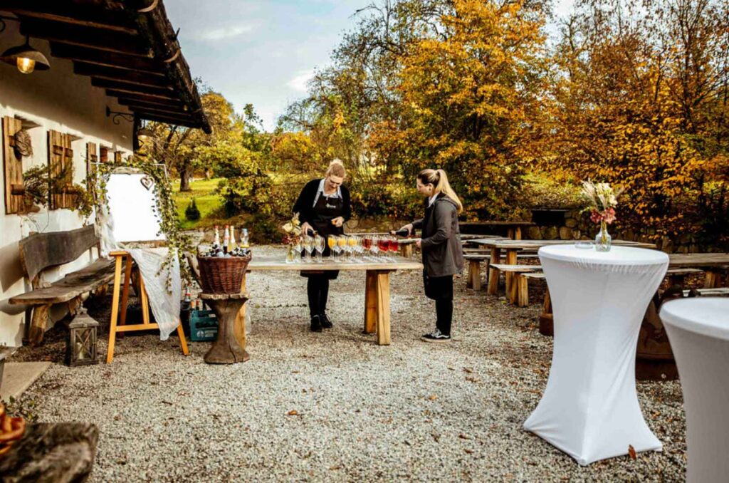Zwei Personen bereiten Getränke an einem Holztisch vor einem rustikalen Gebäude in Passau zu, umgeben von Bänken, herbstlichen Bäumen und Cocktailtischen mit weißen Tüchern. Die Szene fängt den Charme eines Hochzeitscaterings im Garten auf einer Kiesterrasse ein.